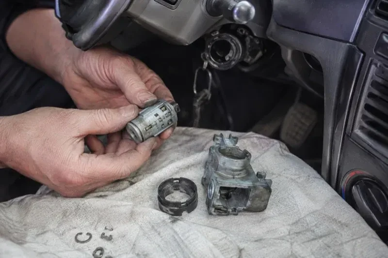 Hands holding a removed ignition cylinder beside a car steering column, with the housing and small parts on a cloth. — Ignition Lock Repair, Kitchener